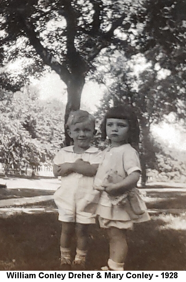 Black and white photo of two very young children, a blond boy in a white onesie and a black-haird girl in a short white dress holding a stuffed animal, standing before a tall tree.